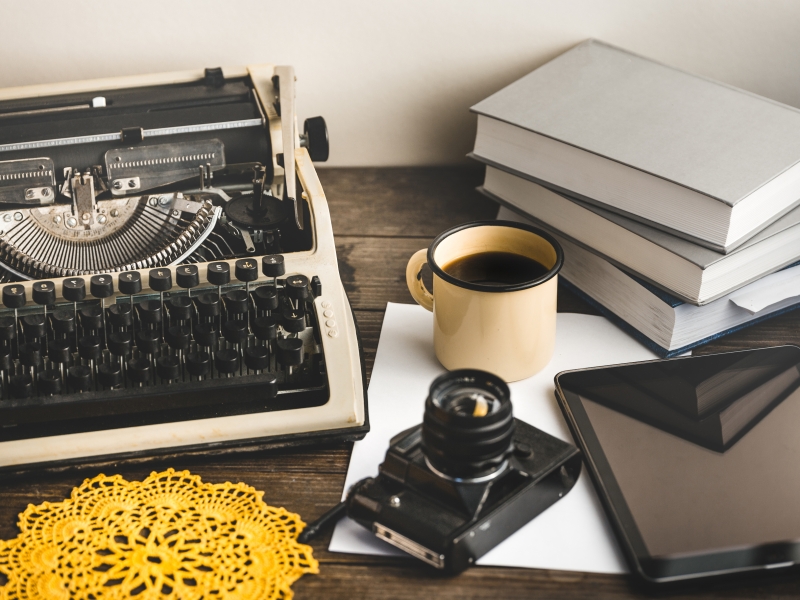 typewriter, stack of books, paper, tablet, camera and paper laid out on a wooden table