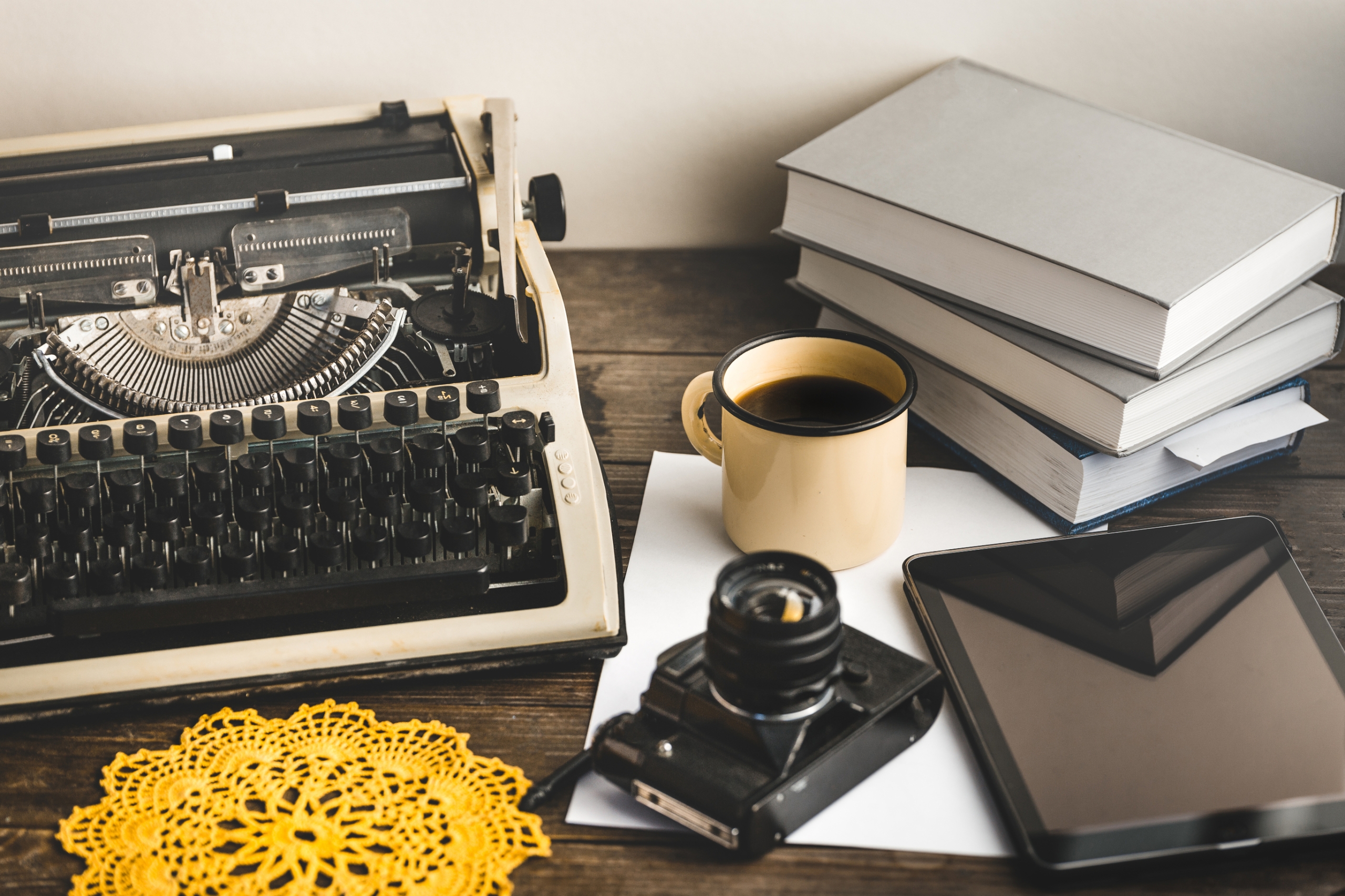 typewriter, stack of books, paper, tablet, camera and paper laid out on a wooden table